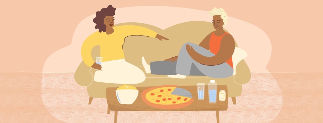 Two women sitting comfortably on a couch with pizza, snacks, and water near them